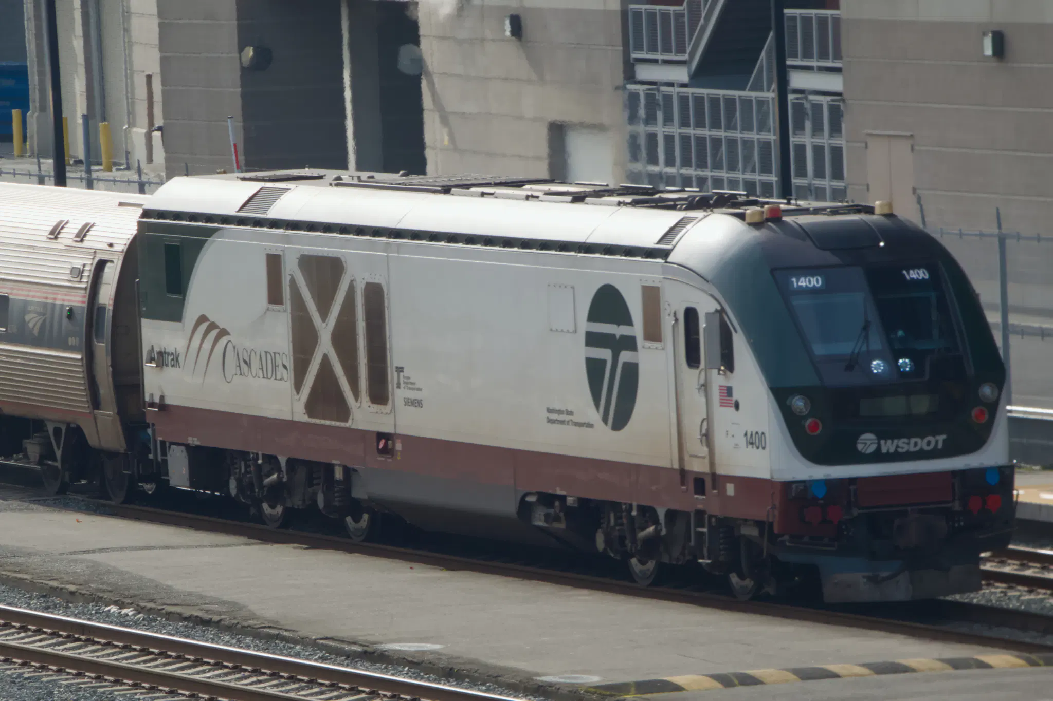 A photo of the lead locomotive of an Amtrak Cascades train, heading north into King Street Station in Seattle. It's a Siemens Charger SC-44 with WSDOT's Cascades livery - the front is green and most of the body is white, with the Cascades logo at the back and a large WSDOT logo on the front just behind the cab. It's pulling some Amfleet cars.