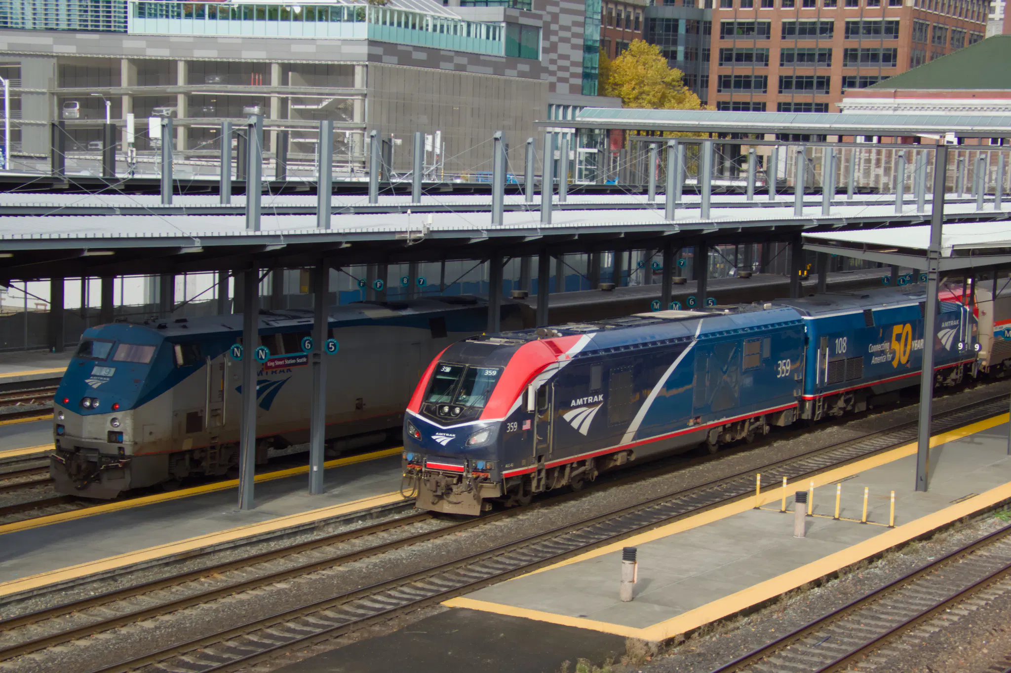 A pair of trains at King Street Station in Seattle. 

The left train, presumably a Cascades train, is lead by locomotive number 19, a 1996 GE P42DC "Genesis" locomotive, with a mostly silver livery with blue on the front and top.

The right train is Coast Starlight, led by number 359, a Siemens Charger ALC-42, coupled to number 108, a 1997 GE P42DC "Genesis".
The Charger has a blue livery, with the front half being a darker blue and hints of red and black, with red next to the window and black above it. The Genesis has a blue livery, with the side saying "Connecting America for 50 YEARS", with the 50 being particularly prominent. There's a bit of red on the front of the cab around the window.
